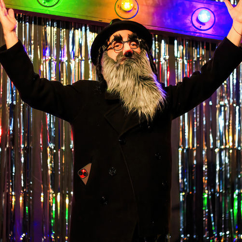 A performer dressed in a black coat, hat, and a long gray beard strikes a dramatic pose on a vibrant stage. The background features shiny metallic curtains and a large wooden frame adorned with multicolored lights, adding a festive and theatrical atmosphere. The individual raises their hands confidently, exuding energy and showmanship.